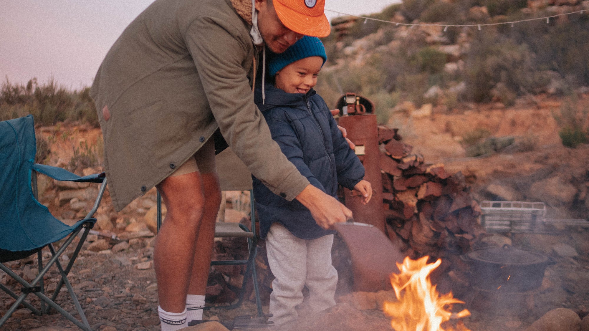 A dad holding his sons hand around the Paddle while fanning it towards the flames outdoors 
