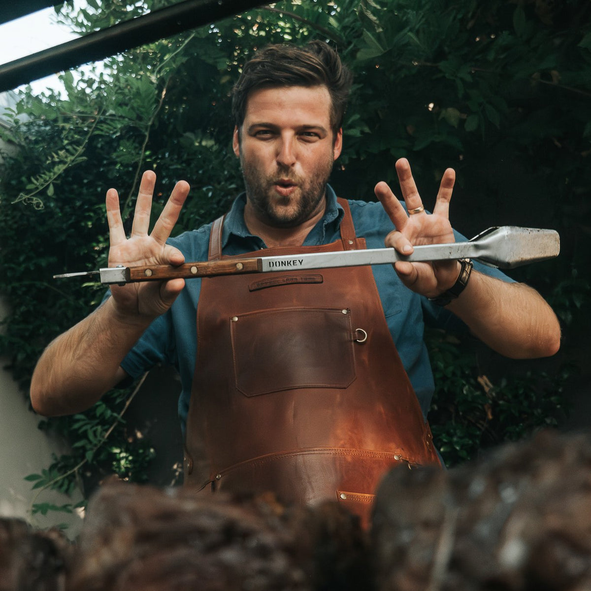 A man wearing a brown leather apron holds a Donkey Tong 69cm up in front of his face, with grilled meat blurred in the foreground and green foliage in the background.